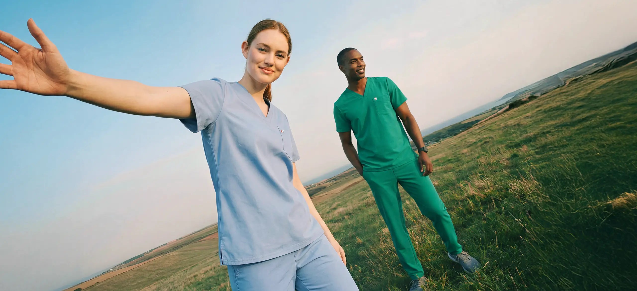 woman wearing blue scrub with left arm outstretched out of left frame standing next to a man in green scrubs. both individuals are standing in a grassy field with a visible blue skyline behind them.